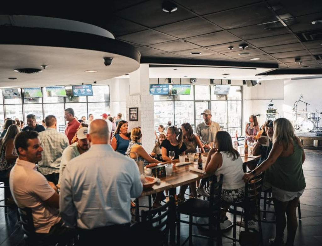 A busy bar scene with people socializing around a large table and at the bar. Multiple TV screens are mounted on the wall, and a drum set is visible in the corner.